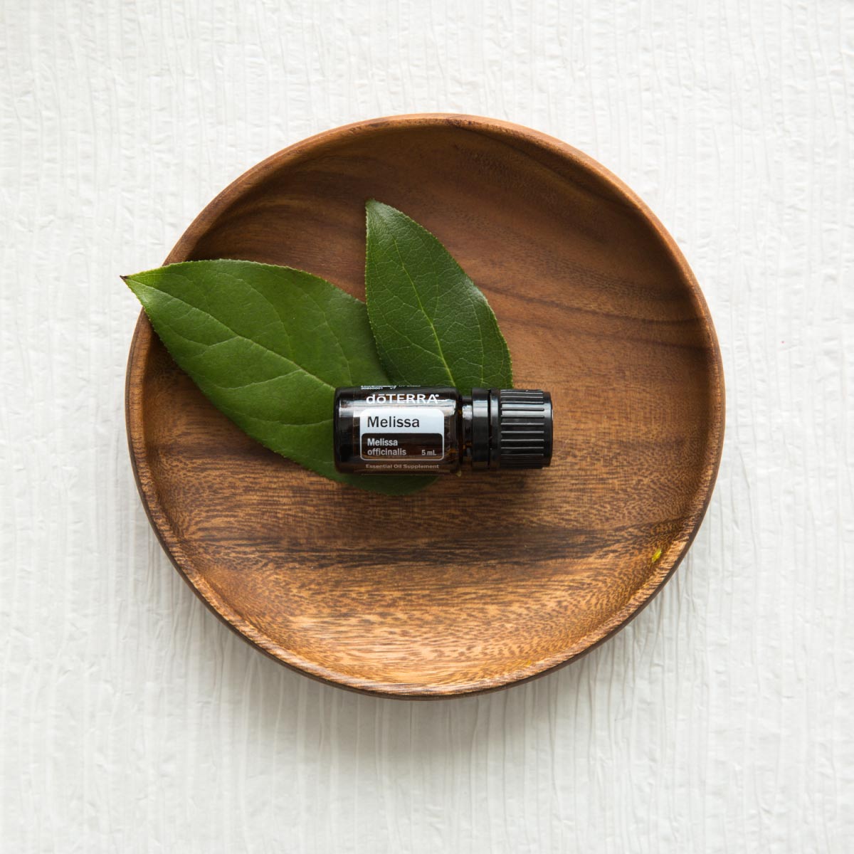 Bottle of Melissa oil resting on a wooden dish, next to green leaves. Melissa oil has many uses and benefits, including benefits for the skin 